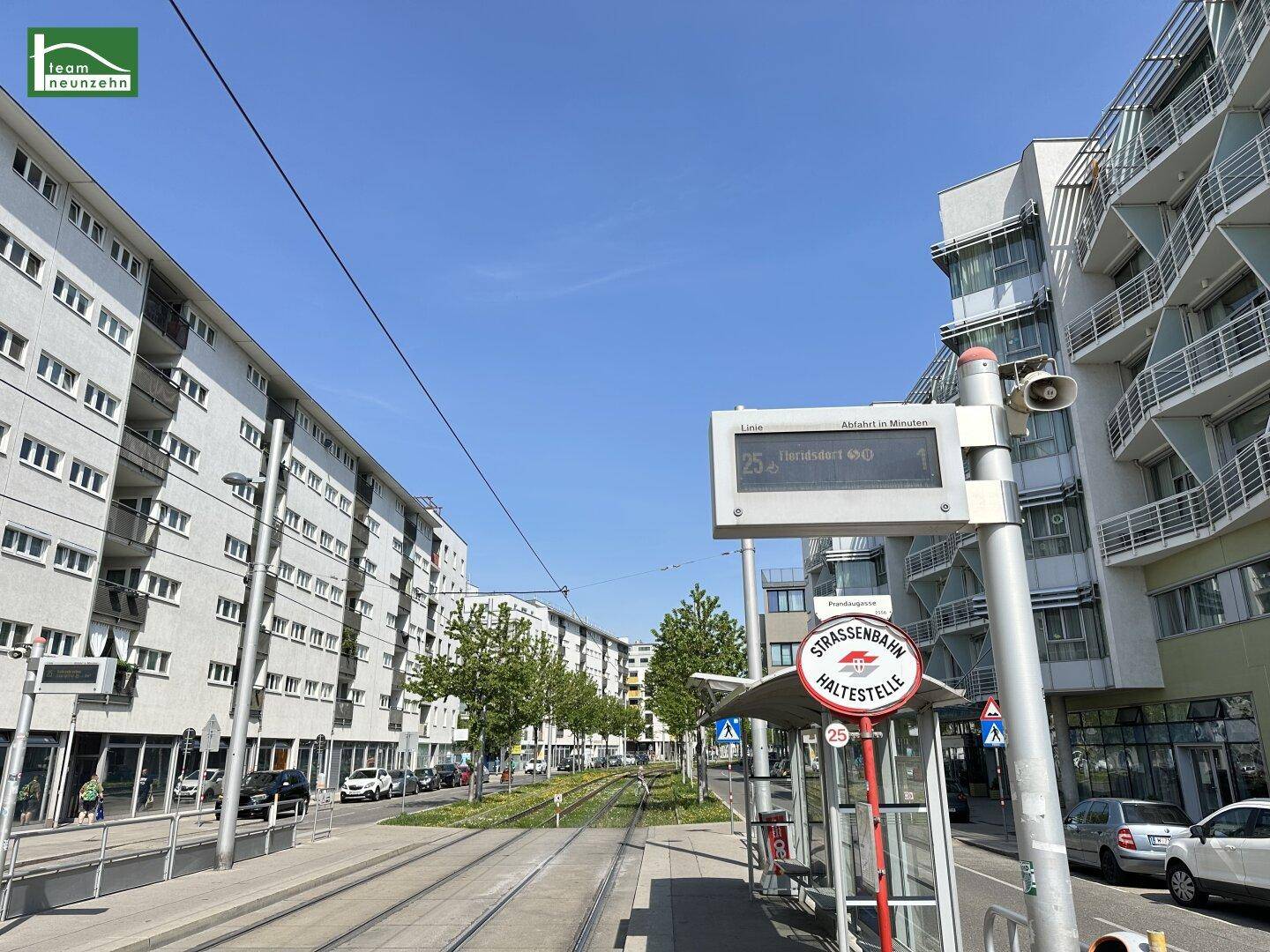 Straßenszene mit Straßenbahnhaltestelle und modernen Wohngebäuden entlang der Gleise.