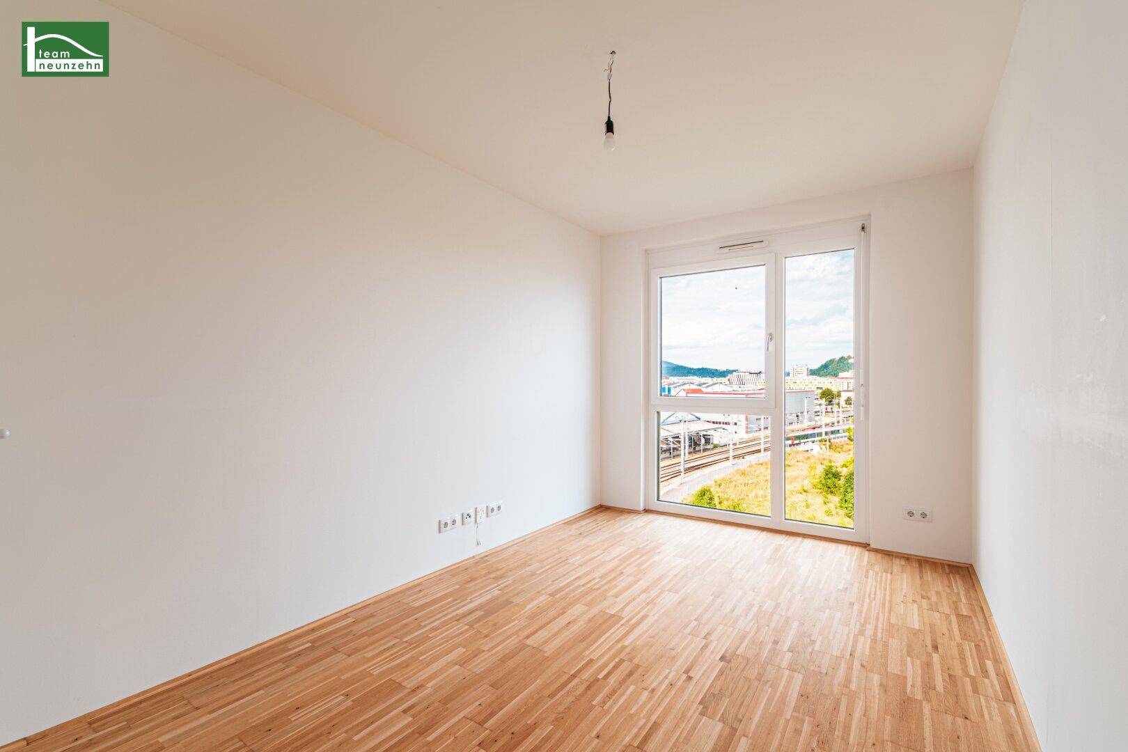 Helles Schlafzimmer mit Holzboden und großem Fenster, das viel Tageslicht hereinlässt.