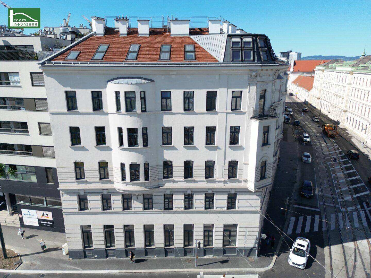 Nähere Luftaufnahme des weißen Gebäudes mit rotem Ziegeldach und Mansarddach in einer belebten Stadtstraße.