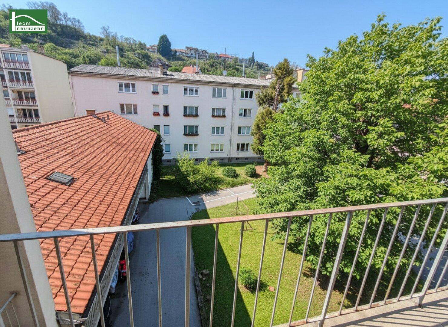 Blick vom Balkon auf einen grünen Innenhof mit Bäumen und umliegenden Wohngebäuden unter blauem Himmel.