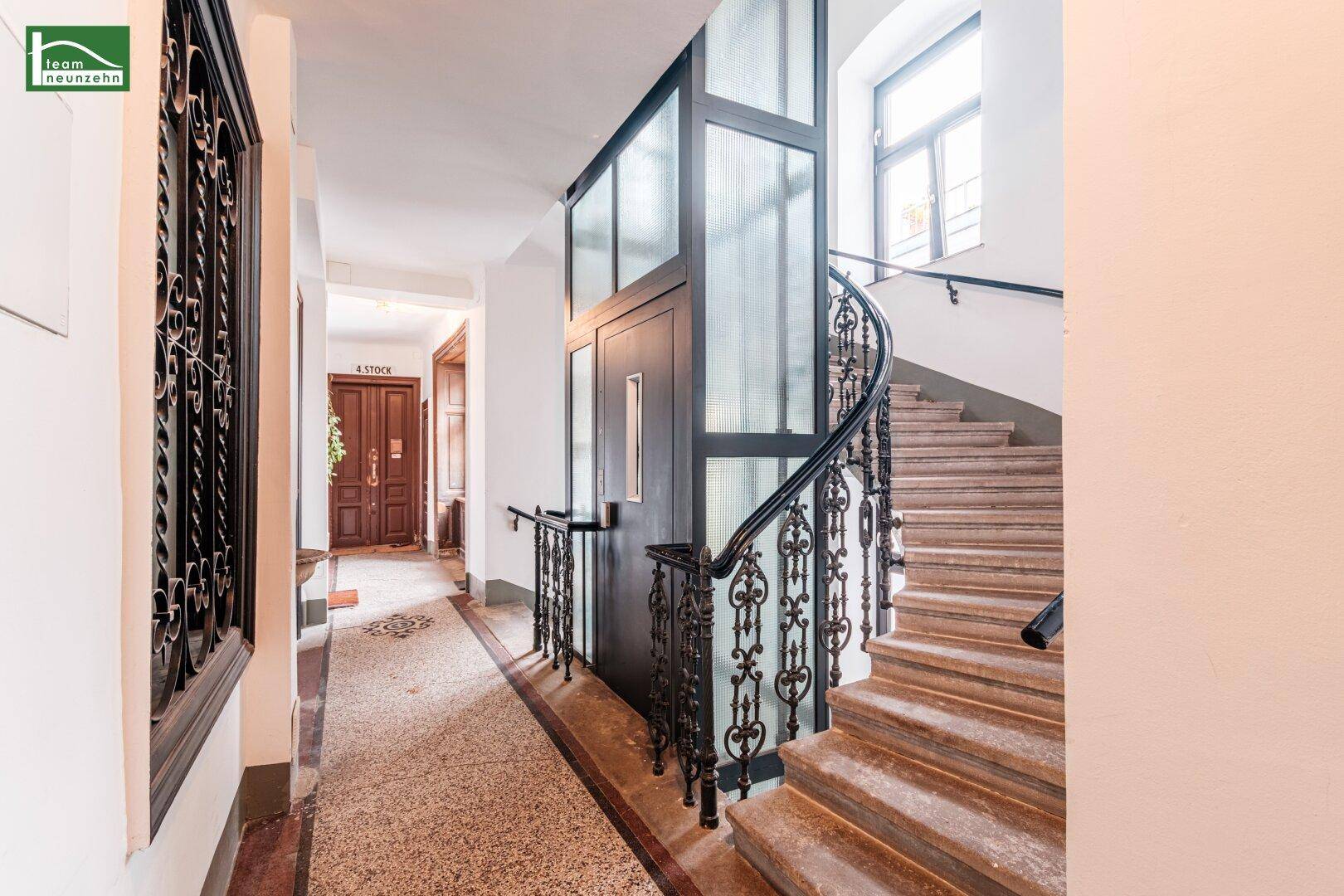 Heller Hausflur mit modernem Glasaufzug, eleganter Wendeltreppe und natürlichem Lichteinfall durch ein großes Fenster.