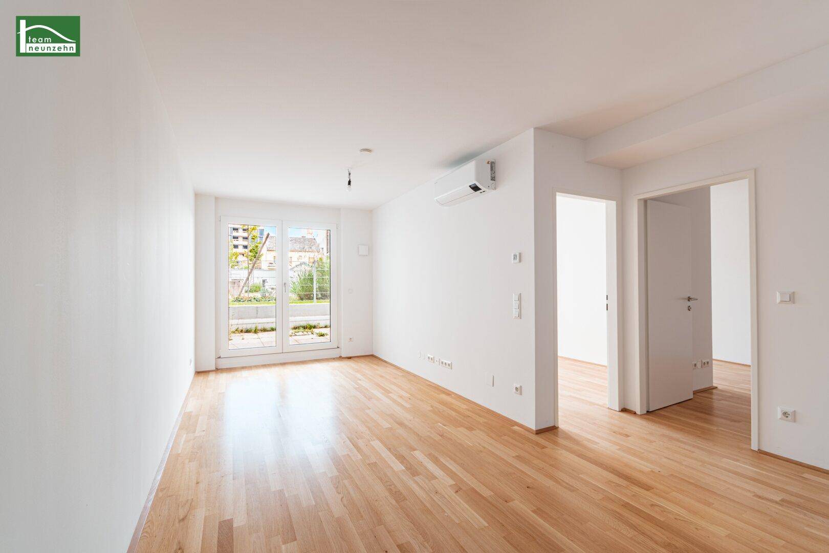 Helles, leeres Zimmer mit Holzboden und großem Fenster, das viel Tageslicht hereinlässt.
