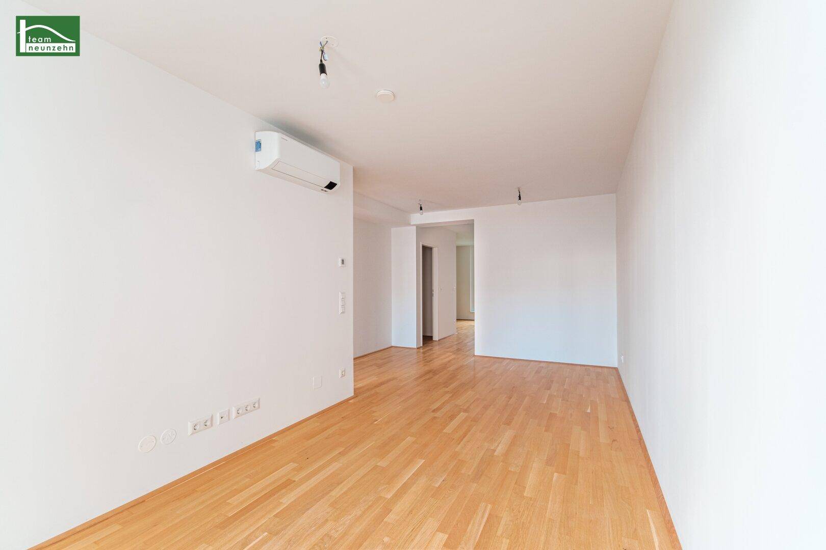 Geräumiges, leeres Zimmer mit hellem Holzboden und einer Klimaanlage an der Wand.