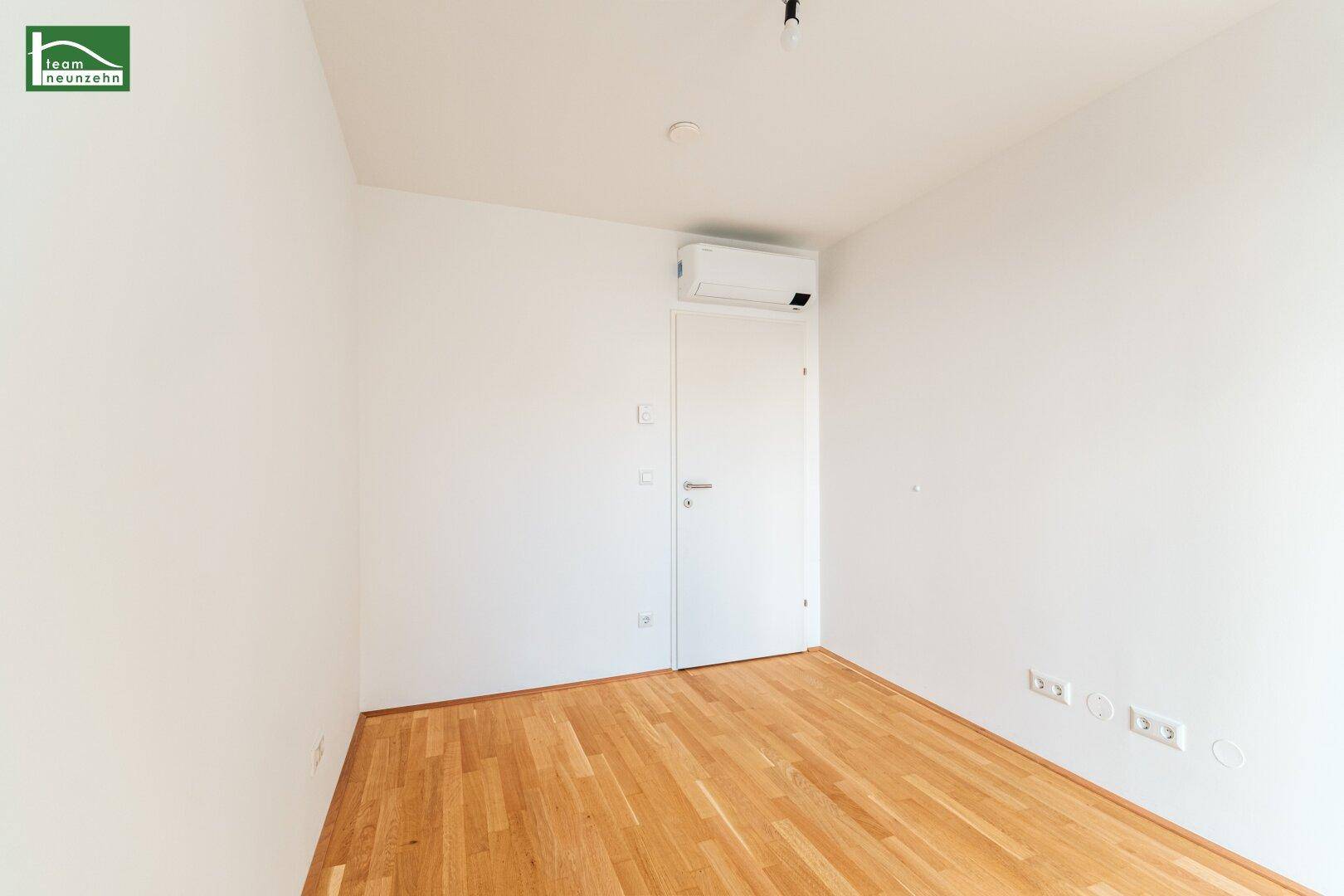 Helles Zimmer mit Holzboden, einer Tür und einer Klimaanlage an der Wand.
