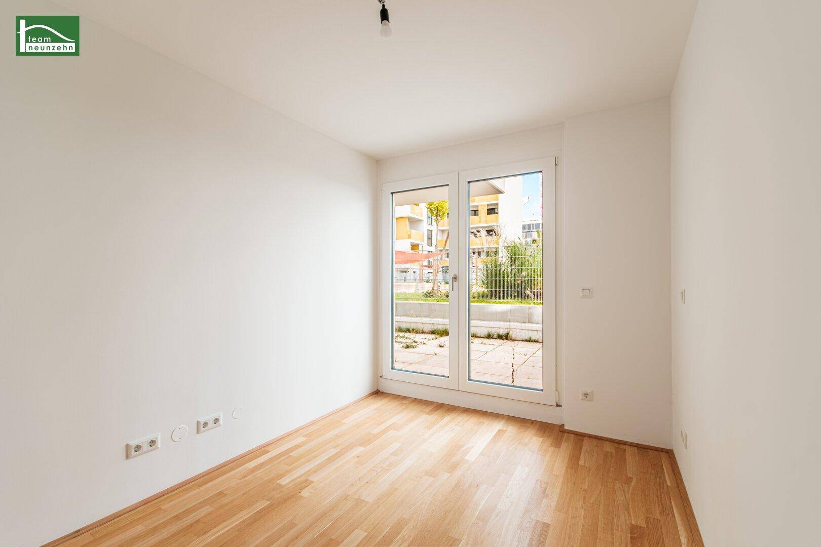 Helles Zimmer mit Holzboden und großem Fenster, das den Blick auf den Außenbereich freigibt.