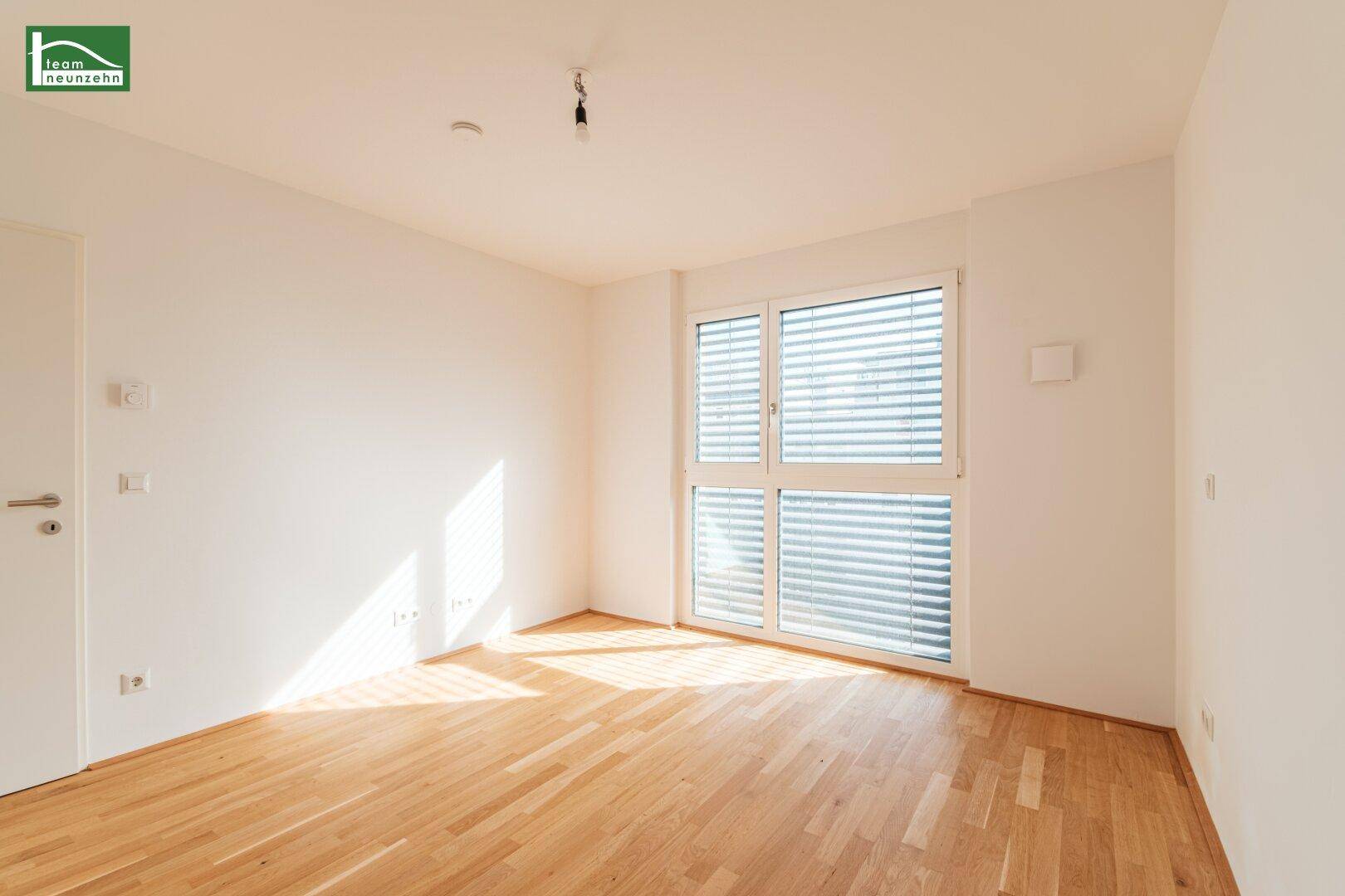 Helles Schlafzimmer mit Parkettboden und großem Fenster mit integrierten Jalousien.