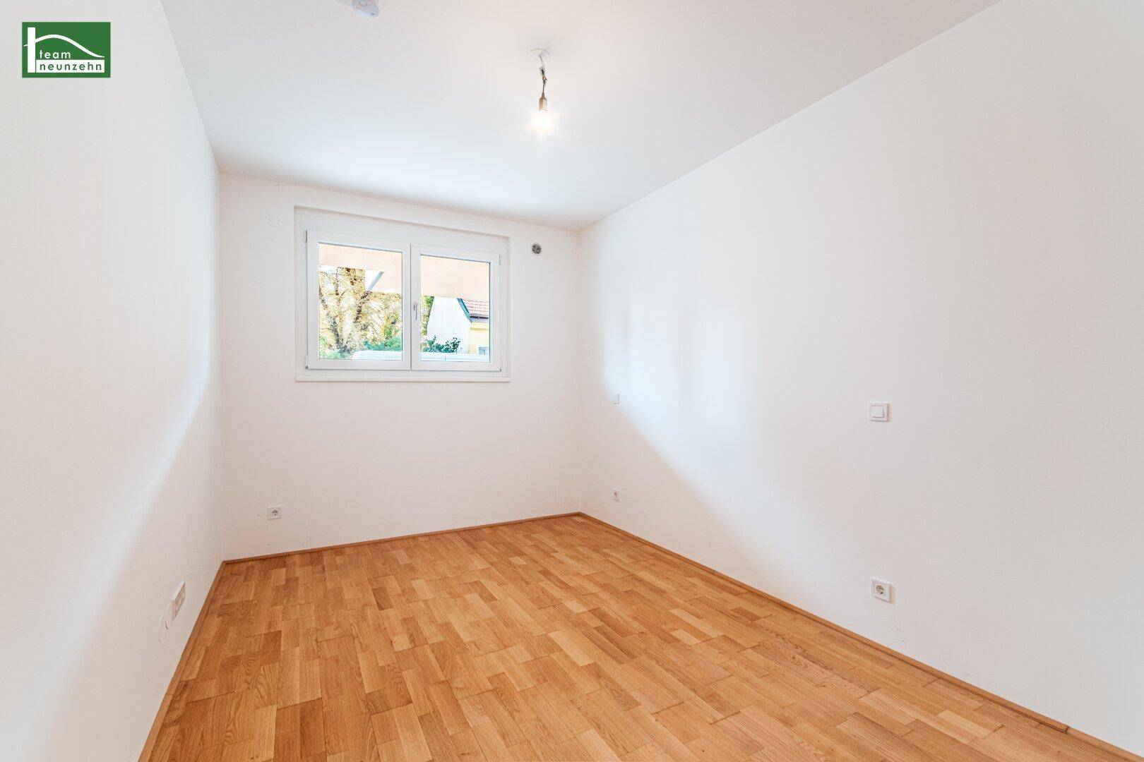 Helles, leeres Schlafzimmer mit Parkettboden und einem Fenster mit Blick ins Grüne.