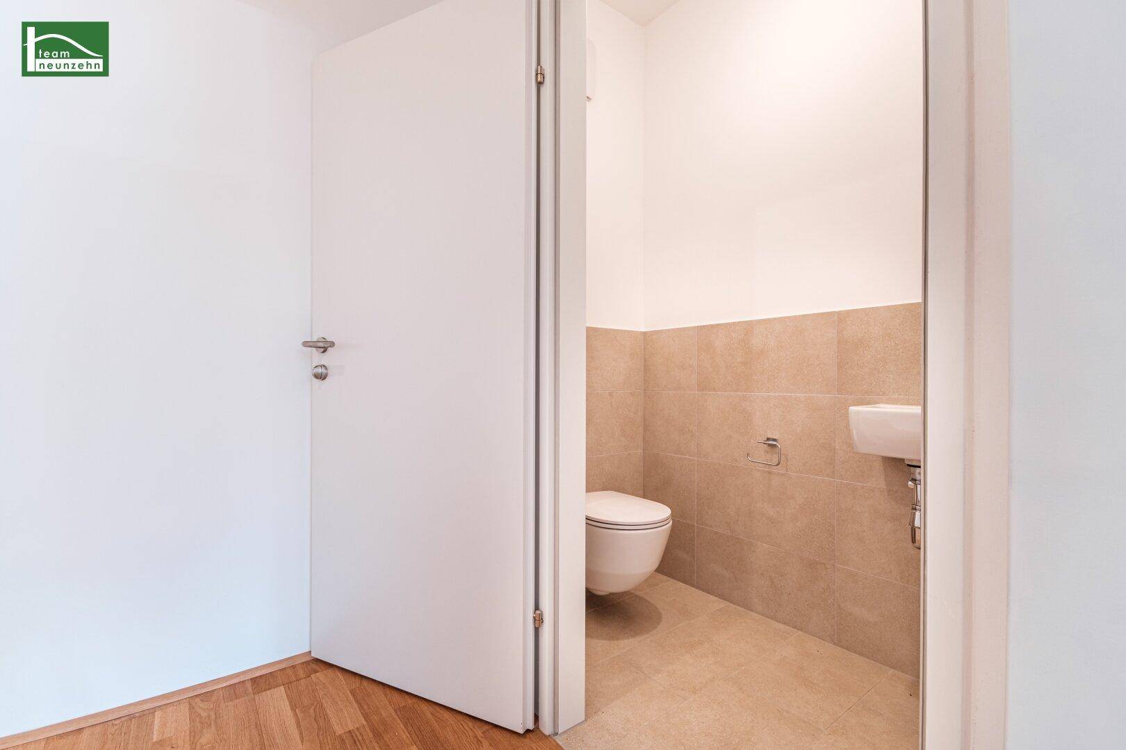 Separates Gäste-WC mit Wand-WC, kleinem Waschbecken und beigefarbenen Fliesen.