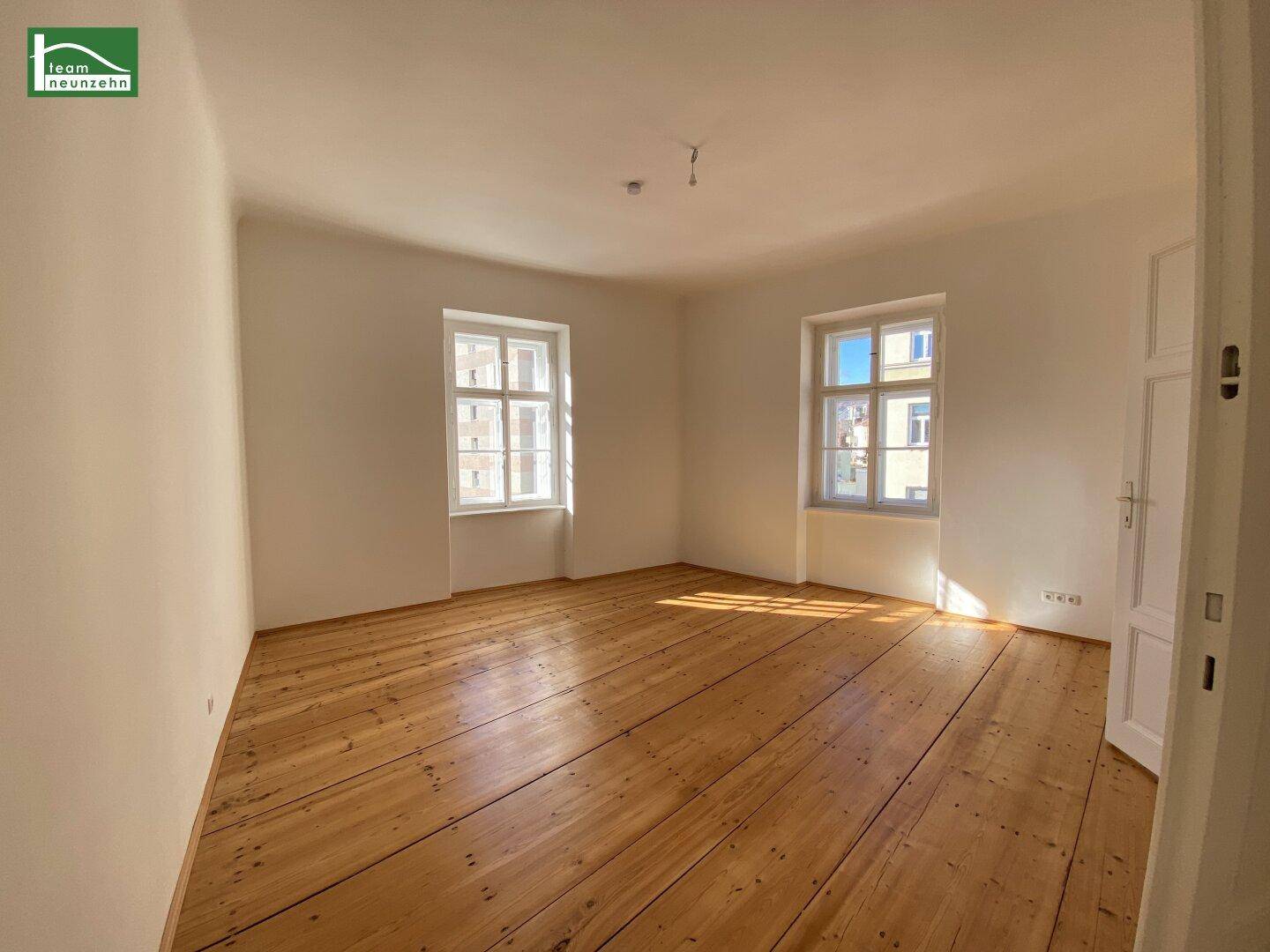 Lichtdurchflutetes Zimmer mit Holzdielenboden und zwei großen Fenstern, ideal als Schlafzimmer.