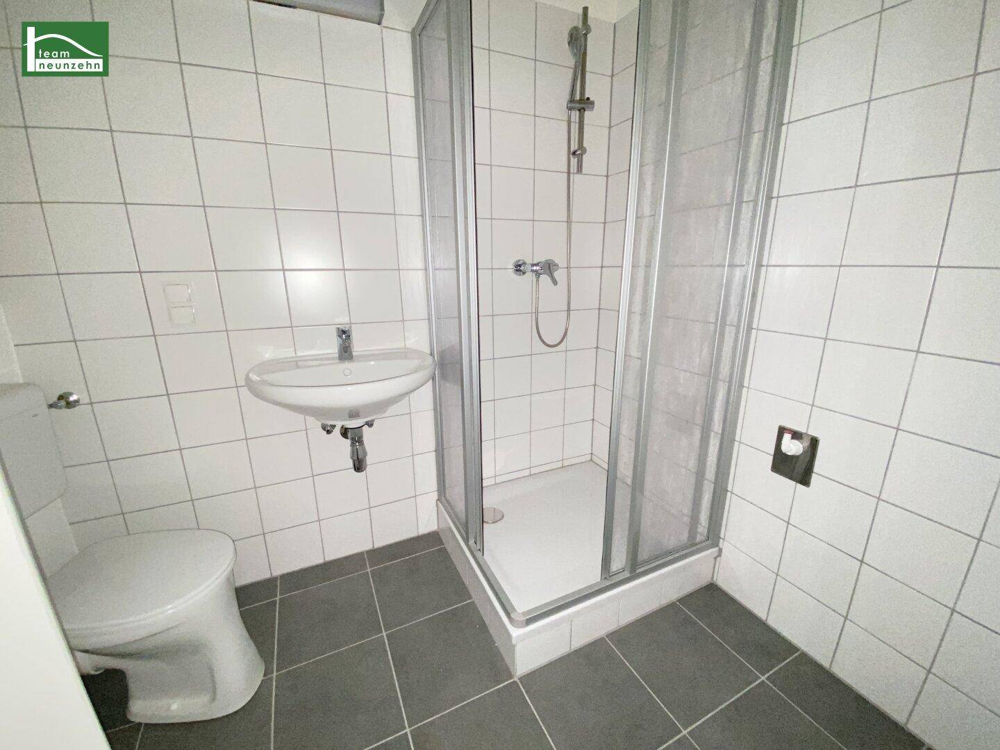 Funktionales Badezimmer mit weißen Wandfliesen, grauen Bodenfliesen, Dusche und WC.