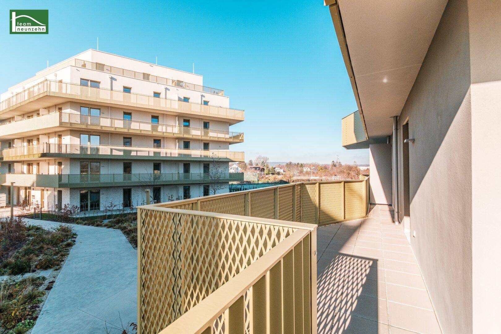 Langer Terrassenbereich mit Gittergeländer und Ausblick auf die Wohnanlage und die Landschaft.