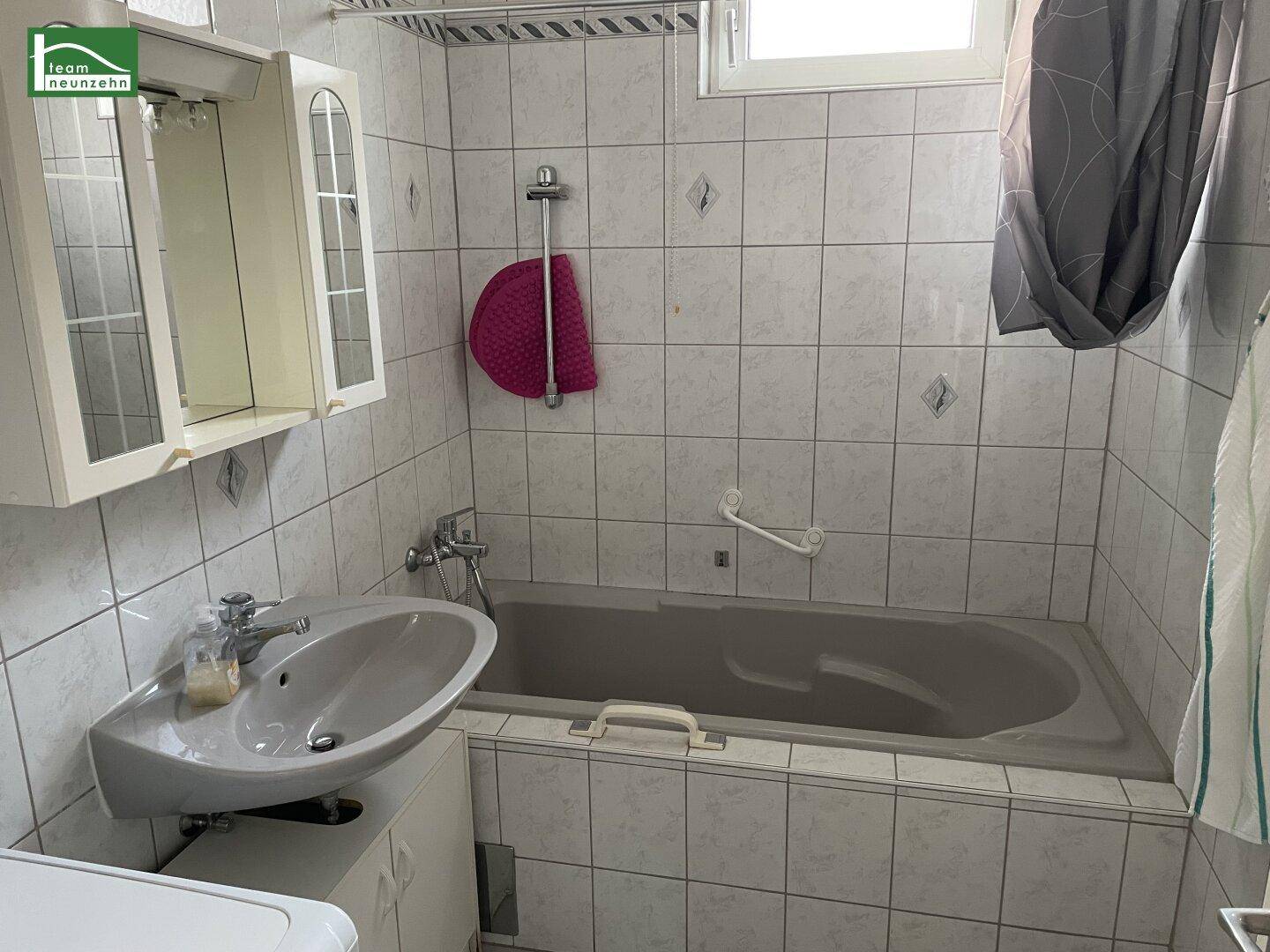 Badezimmer mit hellen Fliesen, Badewanne, Einzelwaschbecken und Fenster.