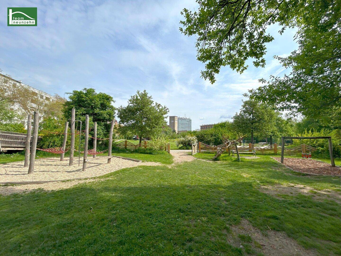 Grüner Parkbereich mit einem natürlichen Kinderspielplatz und hohen Bäumen.