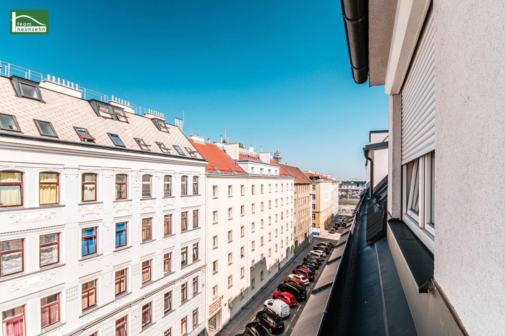 Weitblick über eine belebte Straße mit parkenden Autos und charmanten Altbauten unter blauem Himmel.