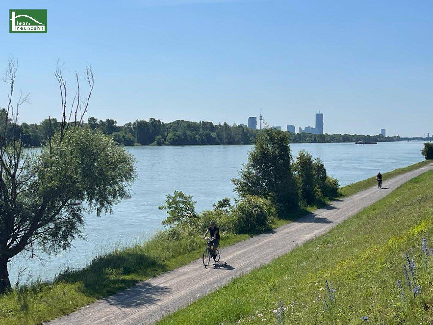 Blick auf den Fluss mit Radweg und Stadtpanorama im Hintergrund an einem sonnigen Tag.