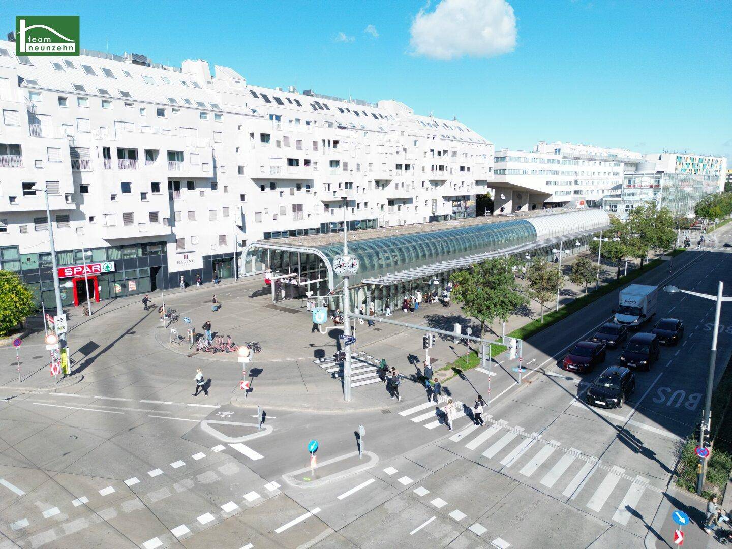 Blick auf eine moderne Stadtkreuzung mit einer U-Bahn-Station und umliegenden Geschäfts- und Wohngebäuden.