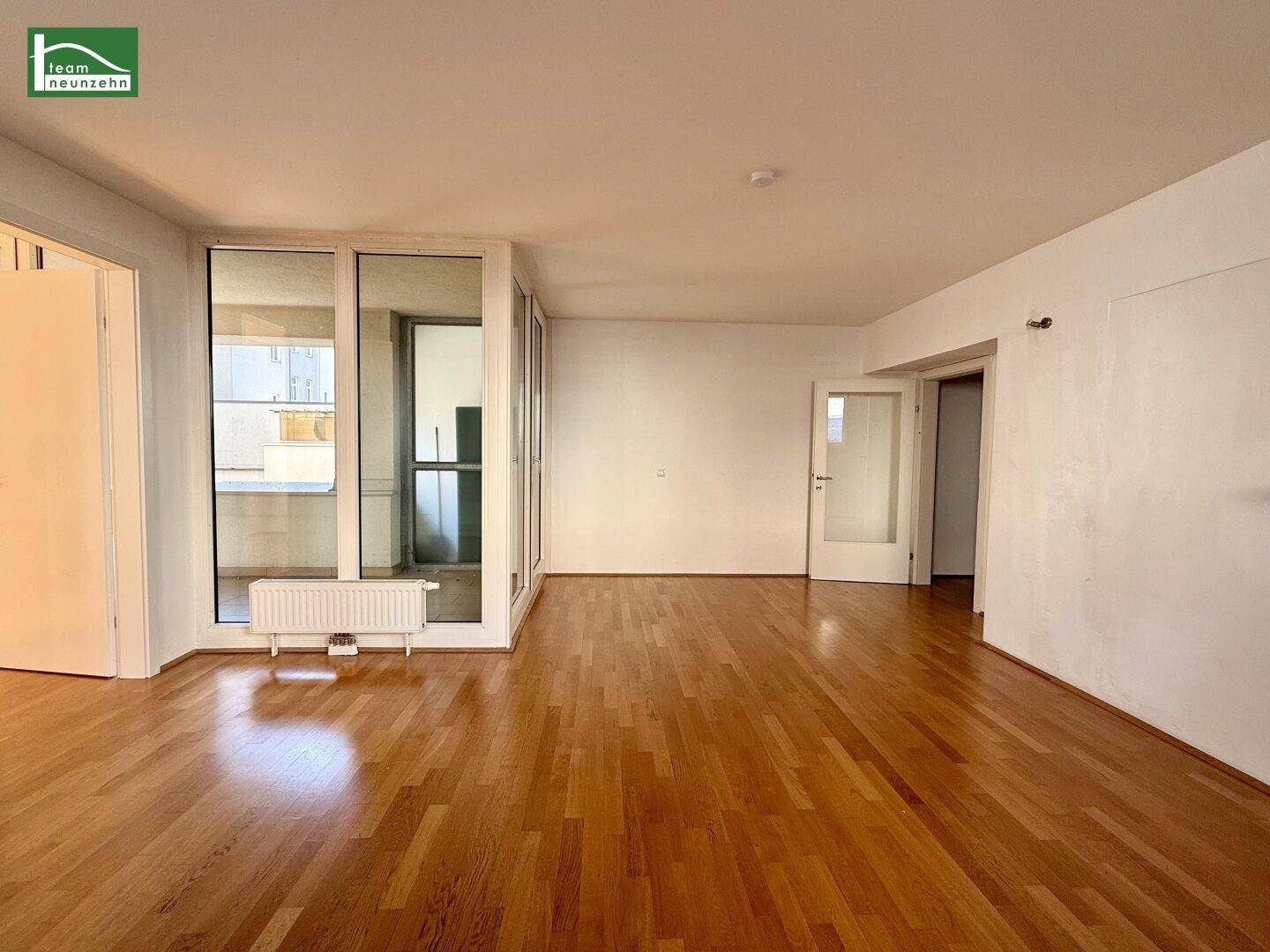 Geräumiges Wohnzimmer mit hellem Parkettboden, großen Fenstern und Zugang zu einem verglasten Balkon.