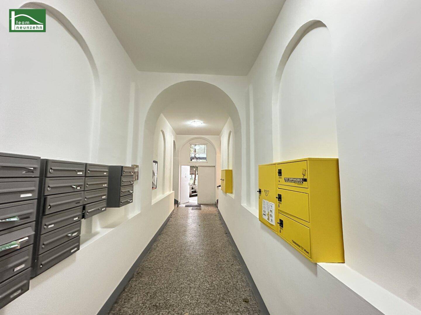 Langer Flur mit Bögen, Briefkästen und einem gelben Postkasten im Eingangsbereich.