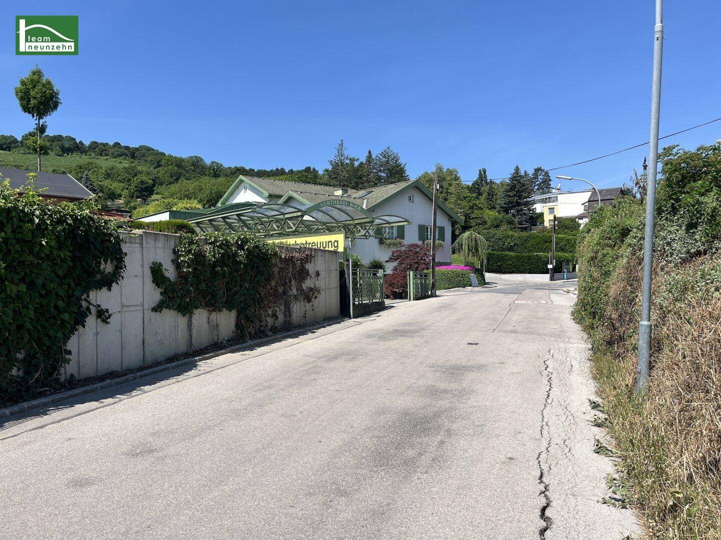 Blick auf die Zufahrt zum Anwesen mit einem Haus, umgeben von üppiger Vegetation an einem sonnigen Tag.