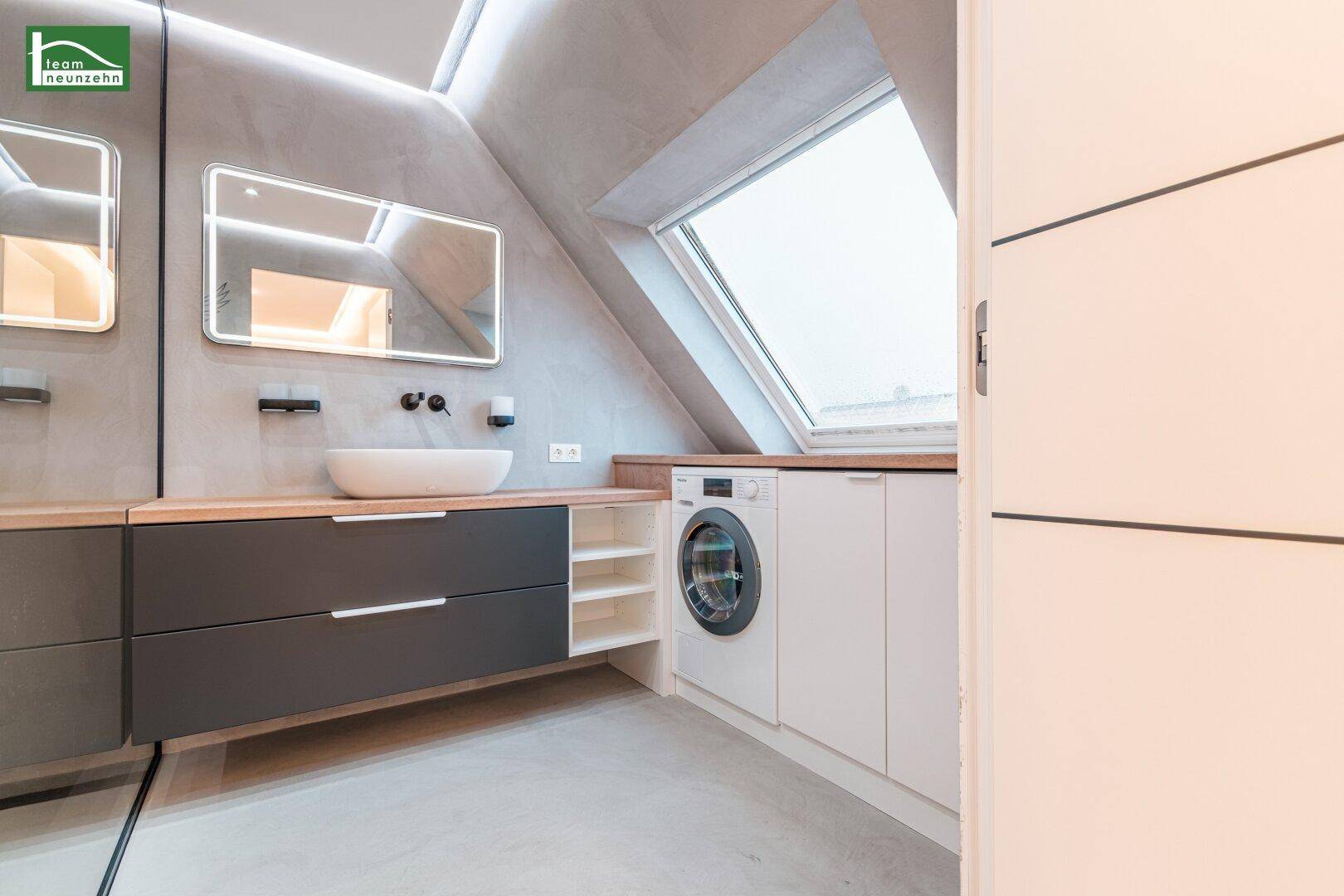 Modernes Badezimmer mit Waschbecken, Spiegel und integrierter Waschmaschine unter dem Dachfenster.