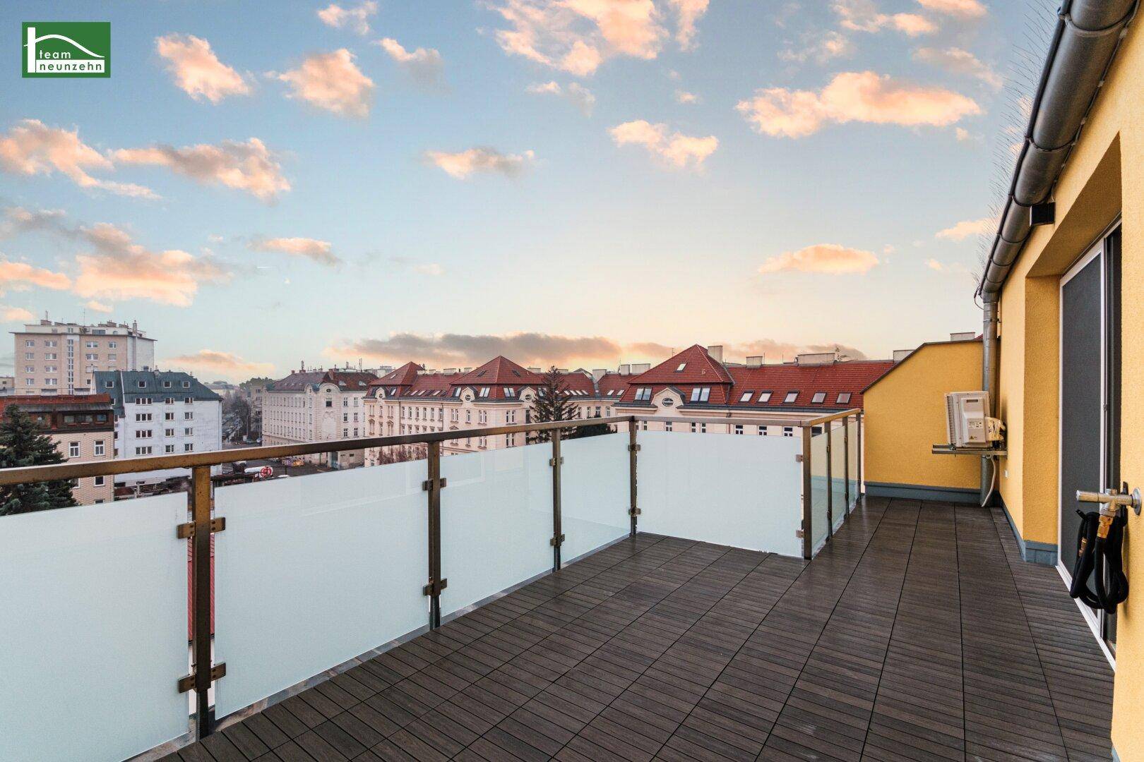 Geräumige Dachterrasse mit Stadtblick, Holzboden und Glasgeländer unter bewölktem Himmel.