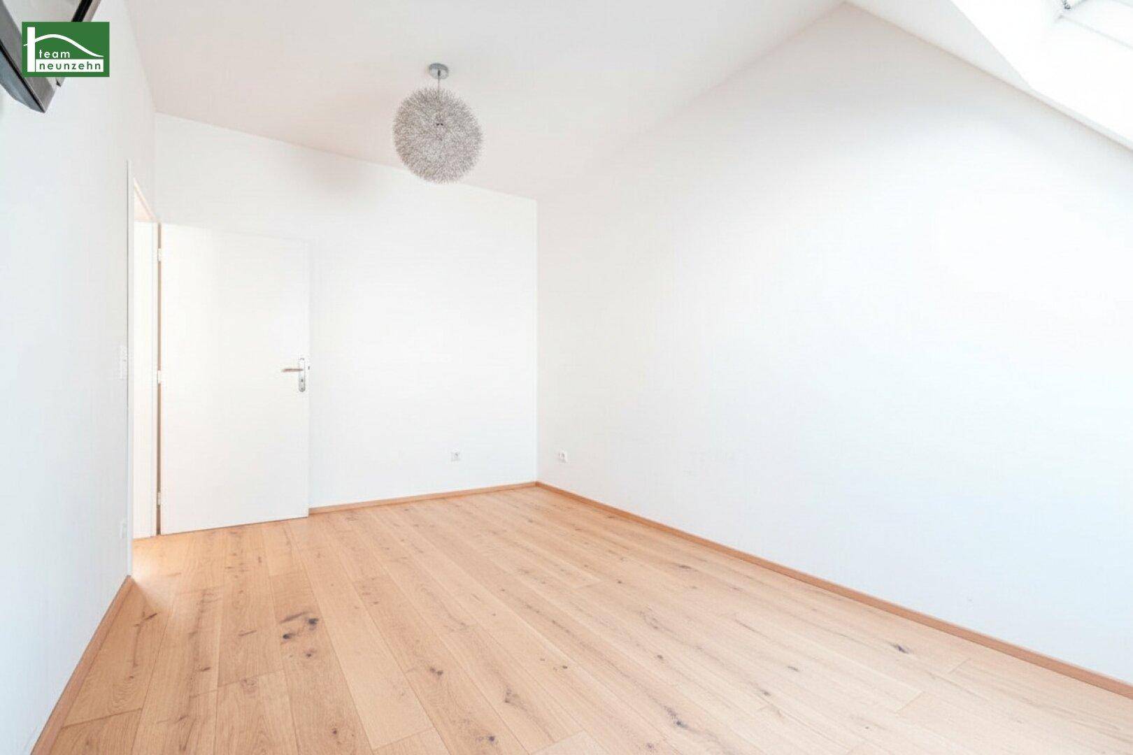 Geräumiger, heller Raum mit Holzboden und weiß gestrichenen Wänden, mit Zugang zu einem weiteren Bereich.