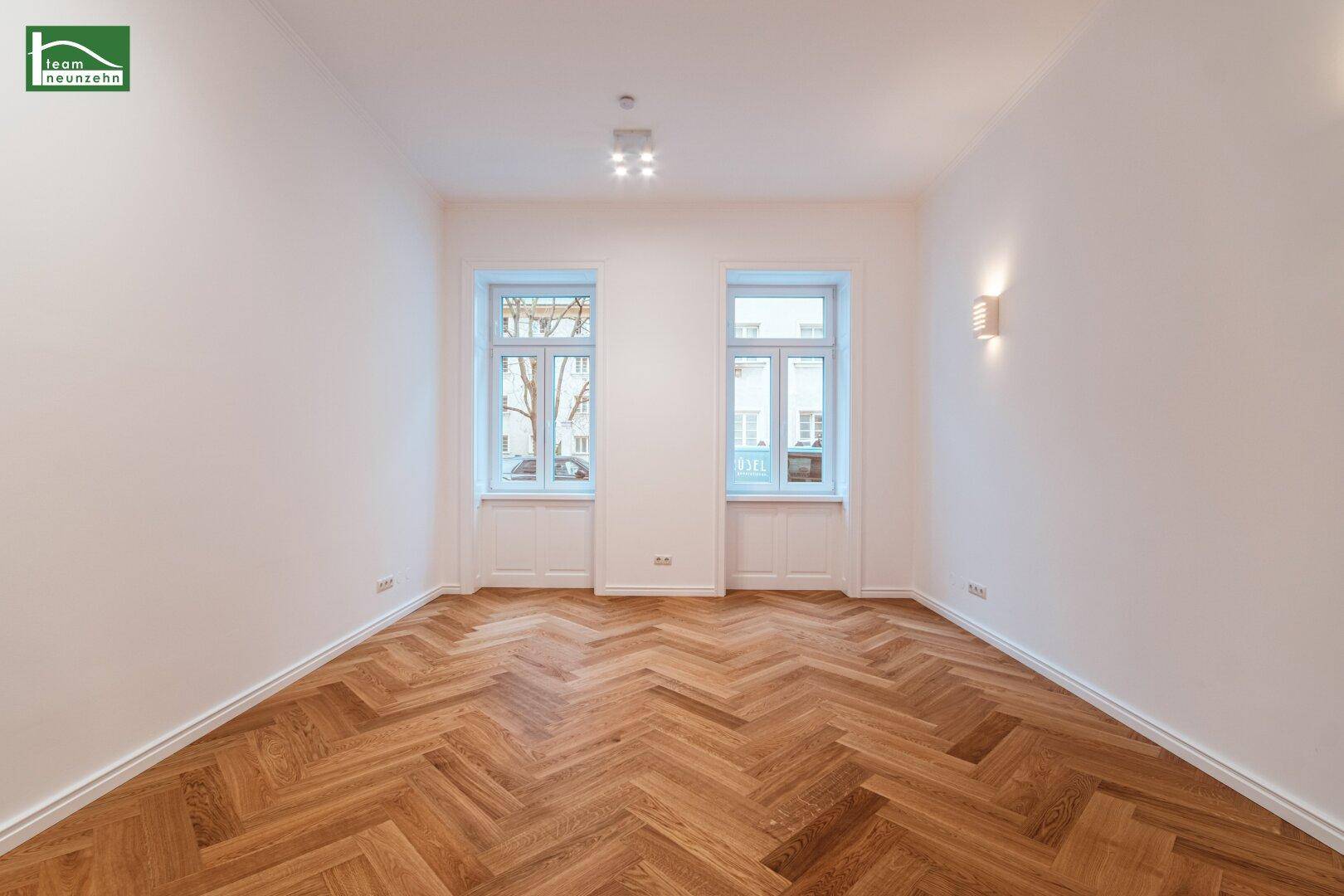 Geräumiges Zimmer mit zwei hohen Fenstern, Fischgrätparkett und Wandbeleuchtung, bietet viel Tageslicht.