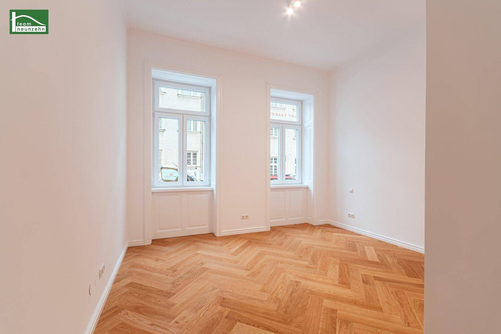 Lichtdurchflutetes Zimmer mit zwei großen Fenstern und klassischem Fischgrätparkett, ideal für Wohnzwecke.