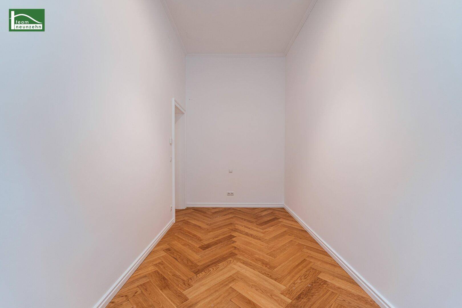 Schmaler, heller Raum mit Fischgrätparkett und einer Tür, ideal als kleines Schlafzimmer oder Büro.
