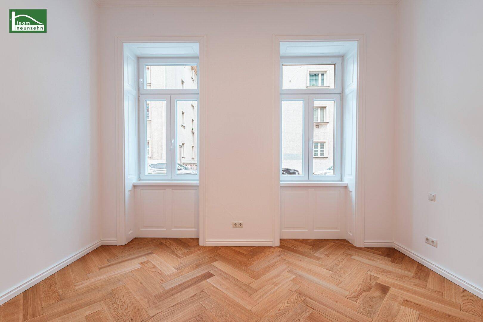 Geräumiges Zimmer mit zwei großen Fenstern und klassischem Fischgrätparkettboden.