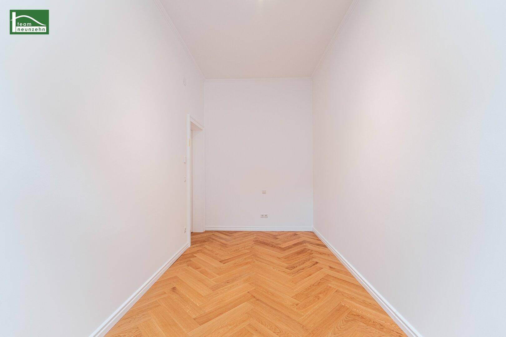 Leerer, schmaler Raum mit weiß gestrichenen Wänden und elegantem Fischgrätparkettboden.