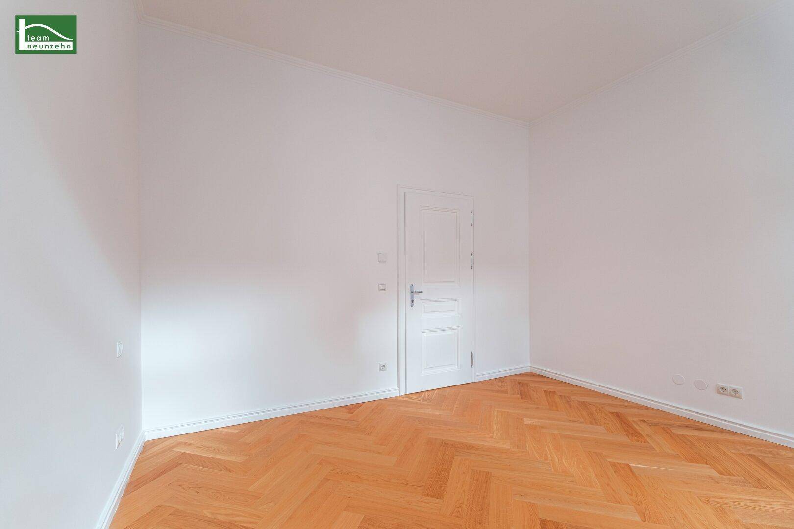 Leerer Raum mit weiß gestrichenen Wänden, klassischer Holztür und Fischgrätparkettboden.