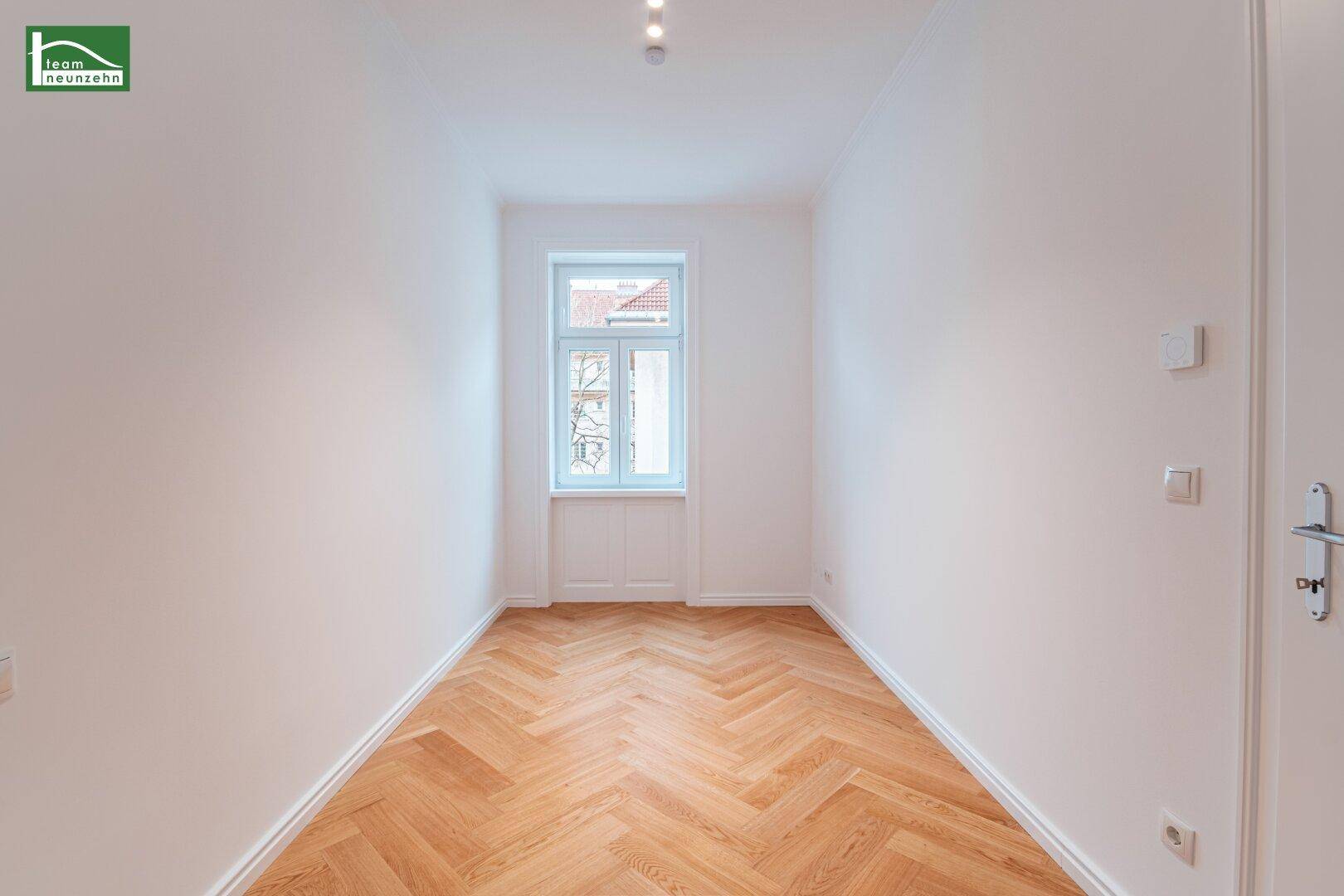 Helles, leeres Zimmer mit Fischgrätparkettboden und einem Fenster, das viel Tageslicht hereinlässt.
