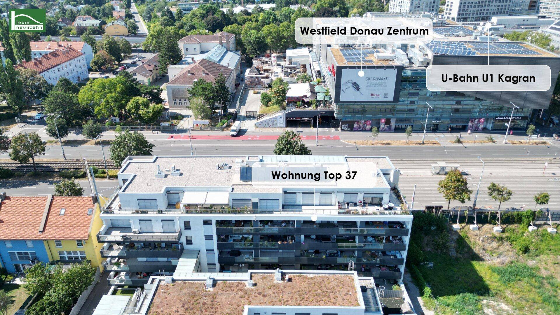 Luftaufnahme eines modernen Wohngebäudes mit Flachdach, umgeben von Grünflächen und städtischer Infrastruktur, inklusive Einkaufszentrum und U-Bahn-Anschluss.