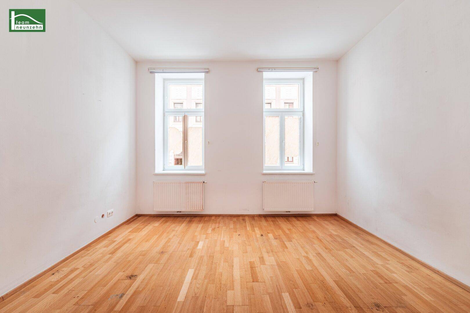 Heller, leerer Raum mit zwei Fenstern, Parkettboden und weißen Wänden.