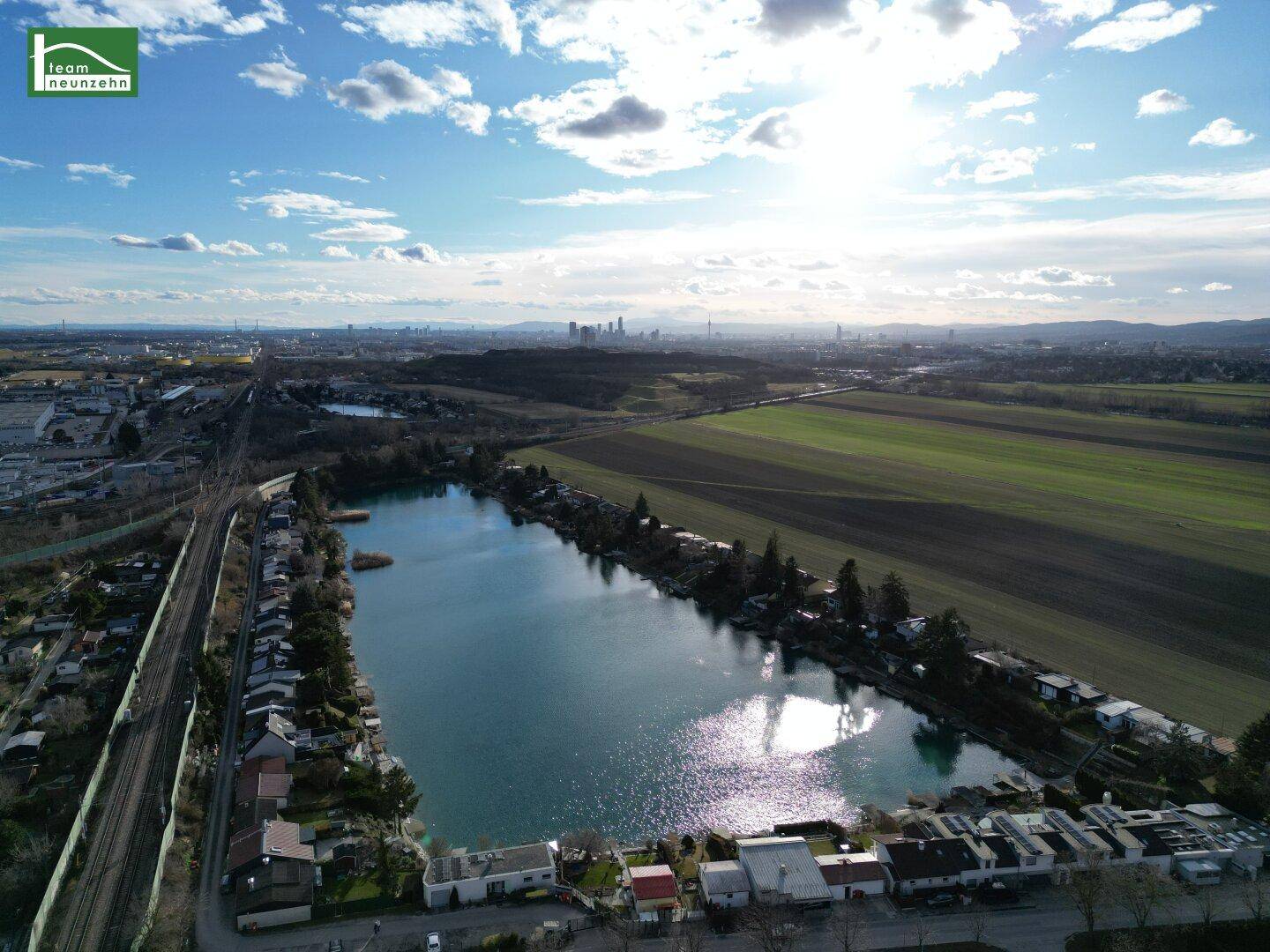 Weitläufige Luftaufnahme mit See, Wohnsiedlung, Feldern und einer fernen Stadtkulisse.