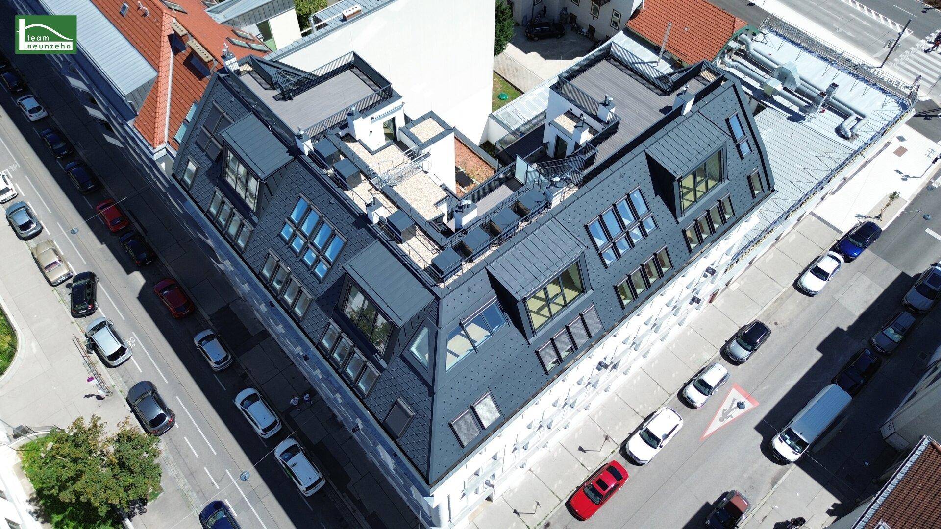 Luftaufnahme des Gebäudedachs mit modernen Dachfenstern und mehreren Dachterrassen in städtischer Umgebung.