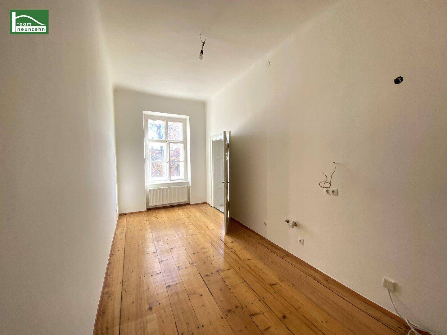 Leerer Raum mit Holzdielenboden, einem Fenster und sichtbaren Elektroinstallationen an der Wand.