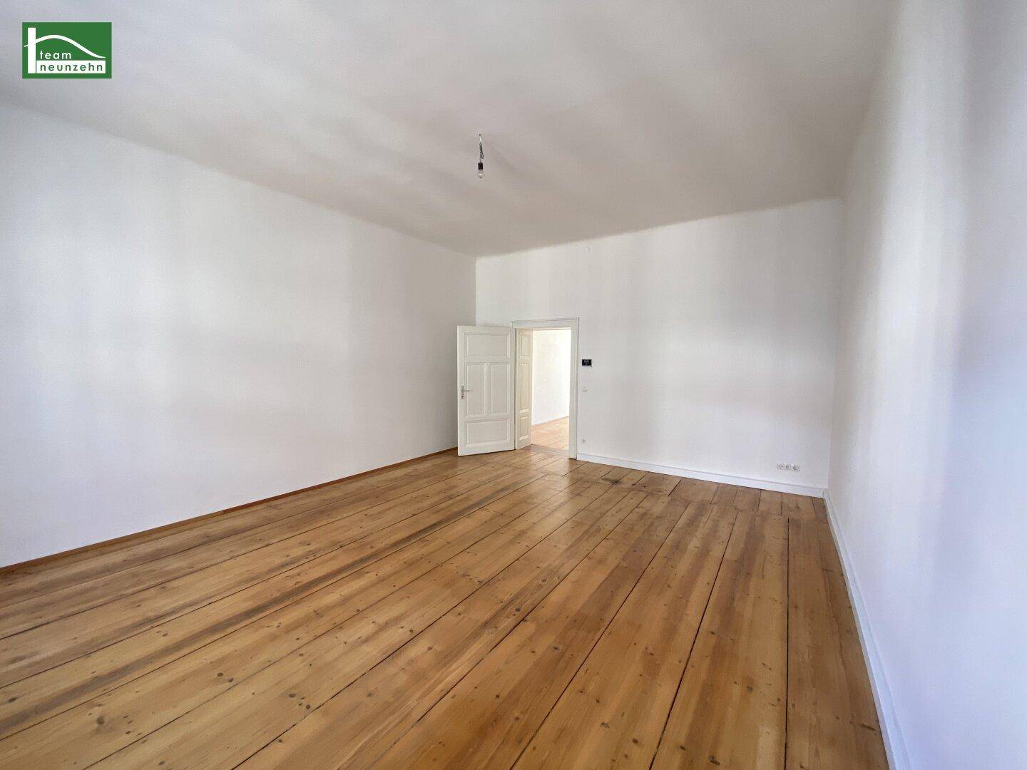 Geräumiges, leeres Zimmer mit Holzdielenboden und einer offenen Tür zu einem weiteren Raum.