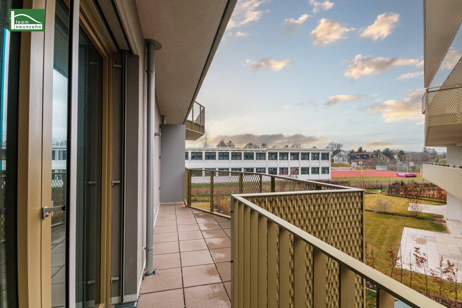 Langer Balkon mit Fliesenboden und Blick auf eine Grünfläche und Sportanlagen.