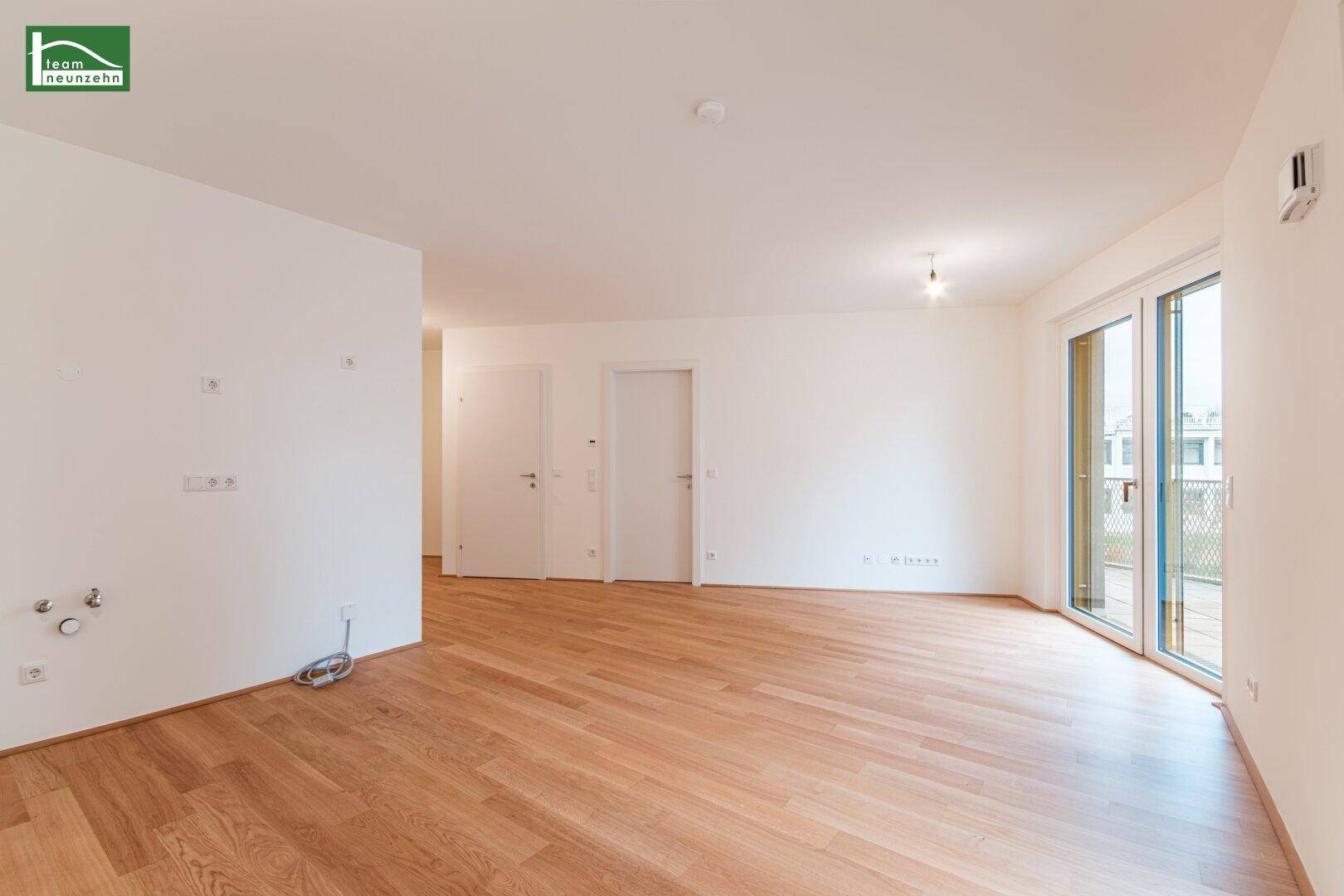 Helles, leeres Wohnzimmer mit Parkettboden und Zugang zu einem Balkon.