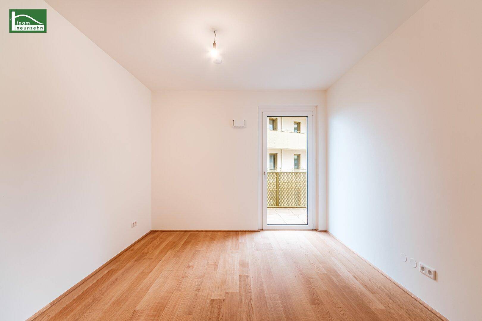 Leeres Zimmer mit Holzboden und Zugang zu einem kleinen Balkon.