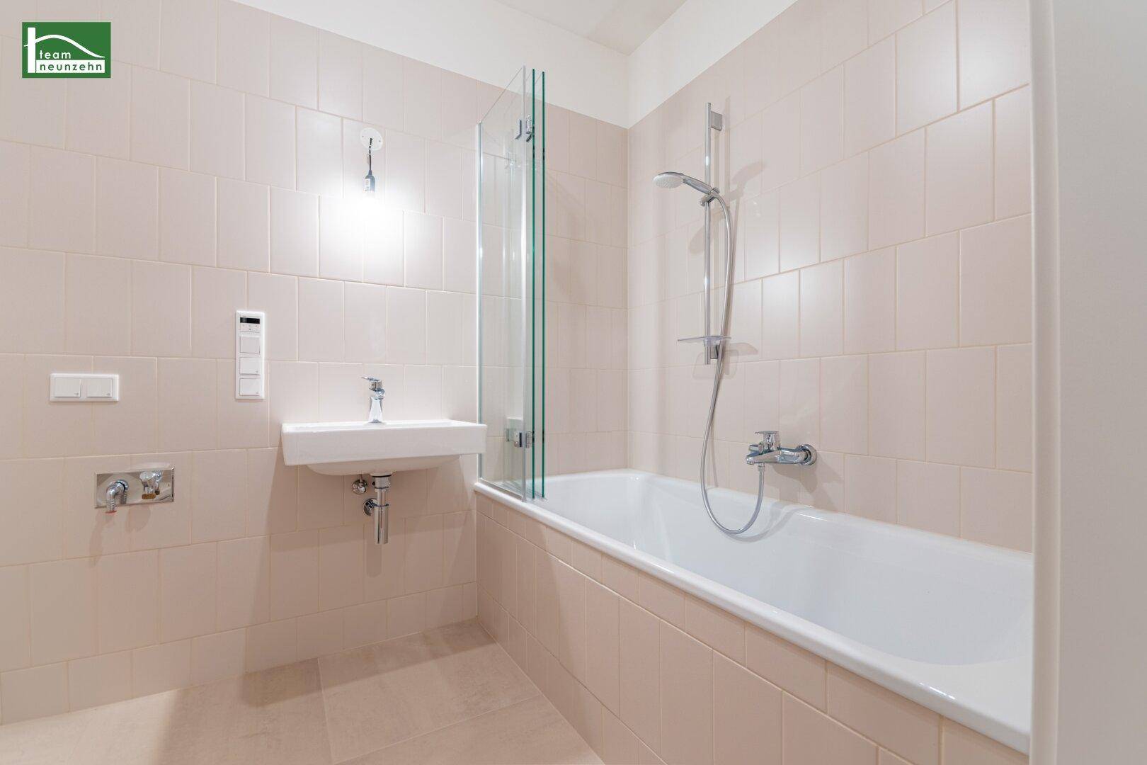 Modernes Badezimmer mit hellbeigen Fliesen, Badewanne mit Duschwand und einem einzelnen Waschbecken.
