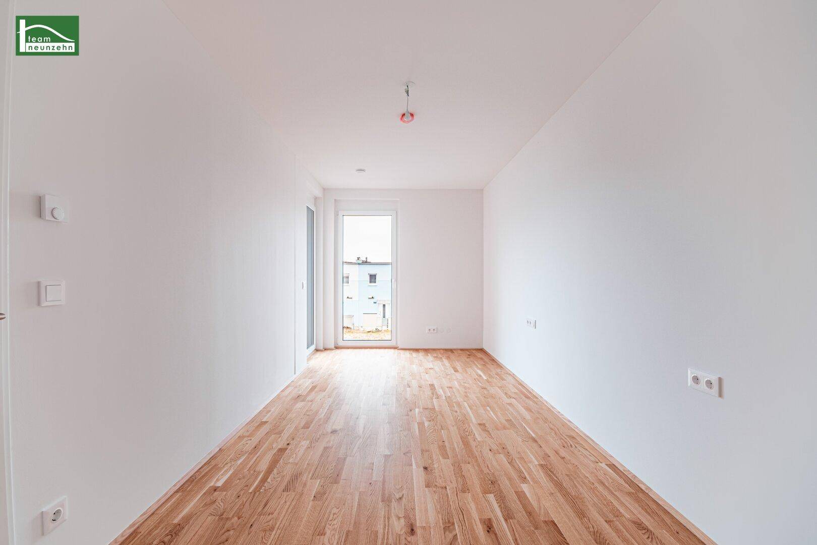 Leeres Zimmer mit hellem Parkettboden und Zugang zu einem kleinen Balkon oder einer Loggia.