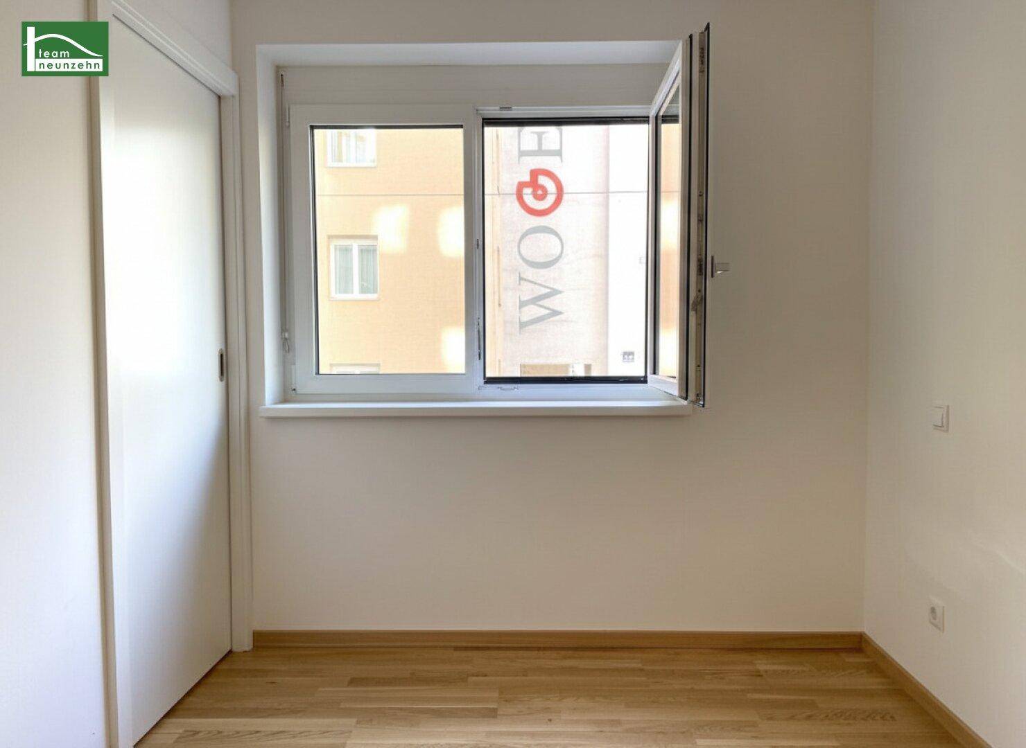 Helles Zimmer mit Parkettboden und einem Fenster, das den Blick auf die umliegenden Gebäude freigibt.