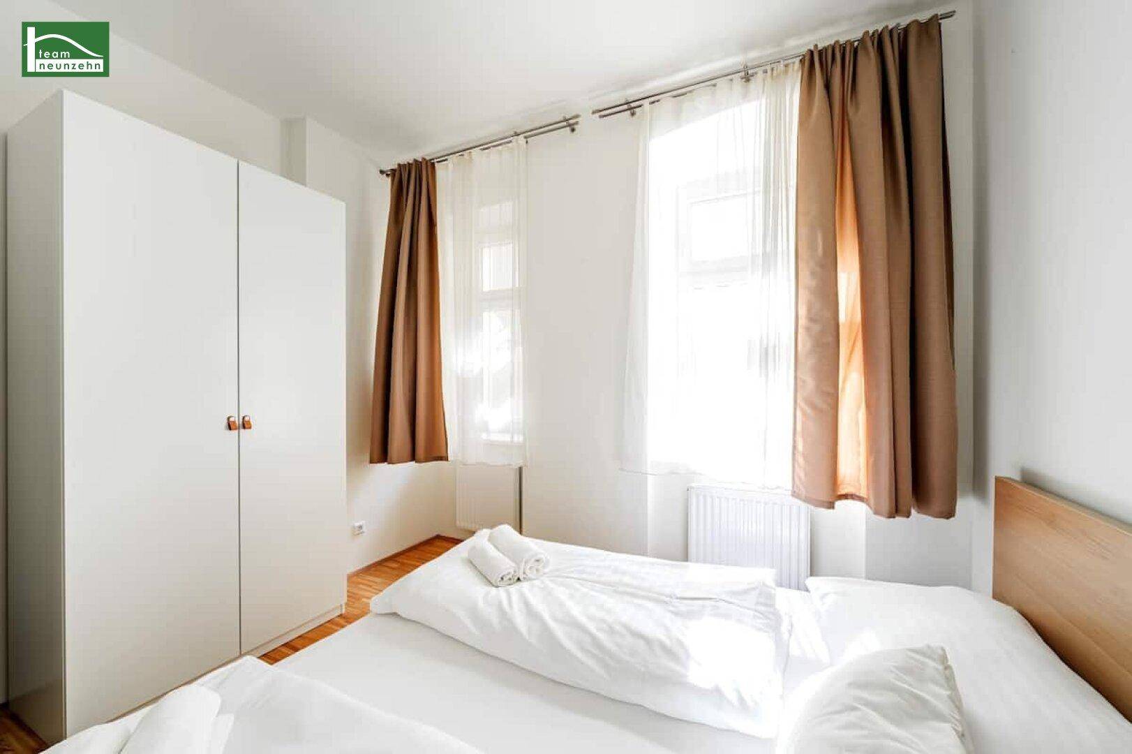 Helles Schlafzimmer mit weißem Kleiderschrank, Holzbett und zwei Fenstern mit braunen Vorhängen.