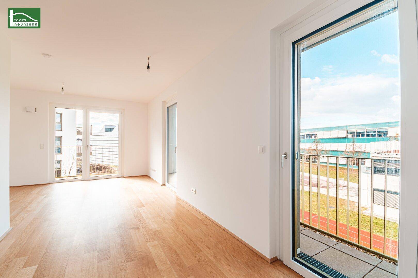Lichtdurchfluteter Raum mit Holzboden und großem Fenster, das auf einen Balkon führt.