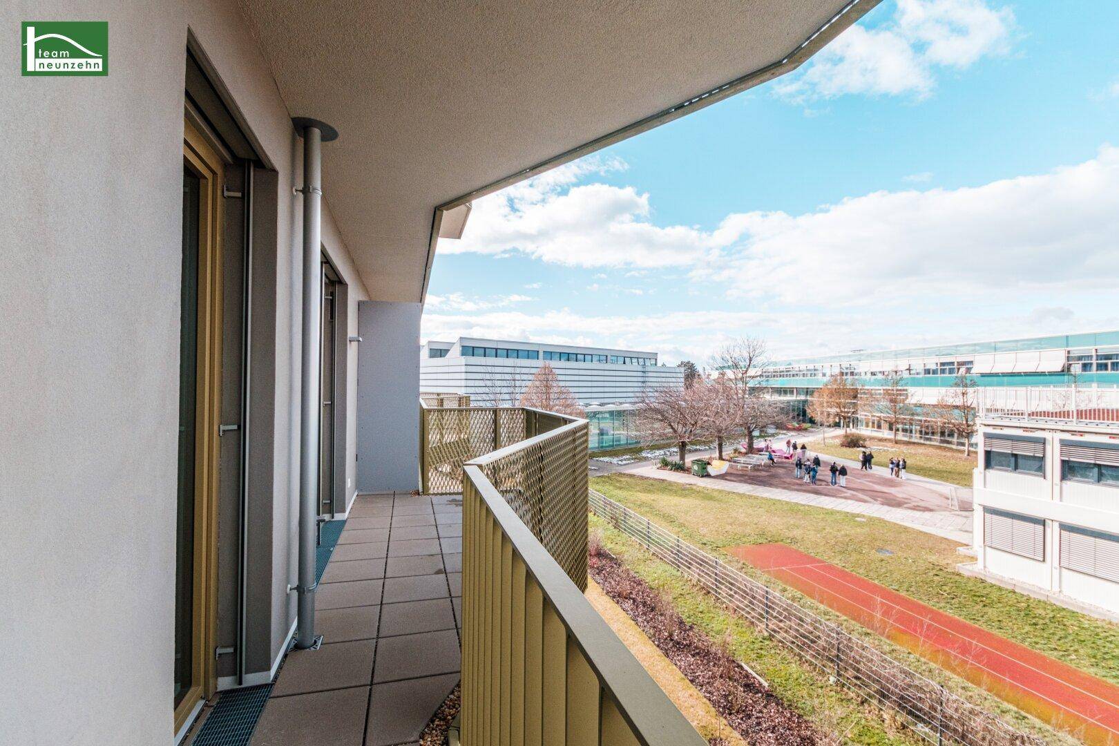 Heller Balkon mit Blick auf eine Sportanlage und grüne Flächen unter blauem Himmel.