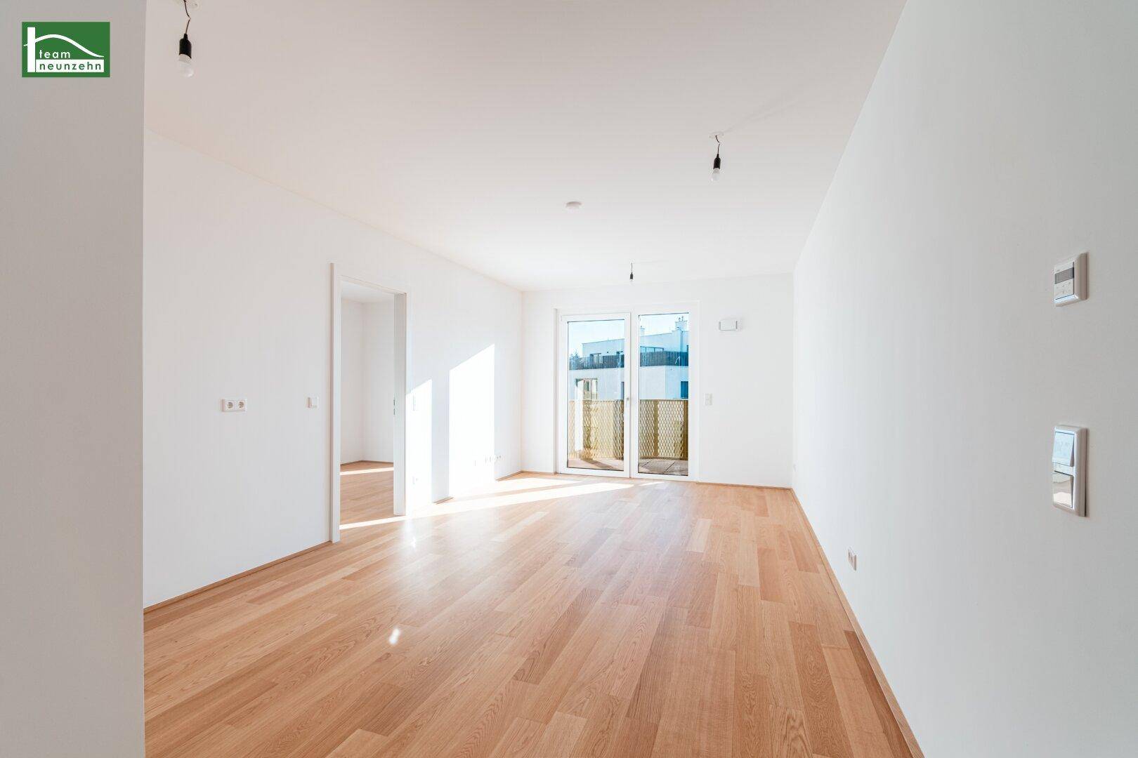 Helles, leeres Wohnzimmer mit hellem Holzparkett und Zugang zu einem Balkon.