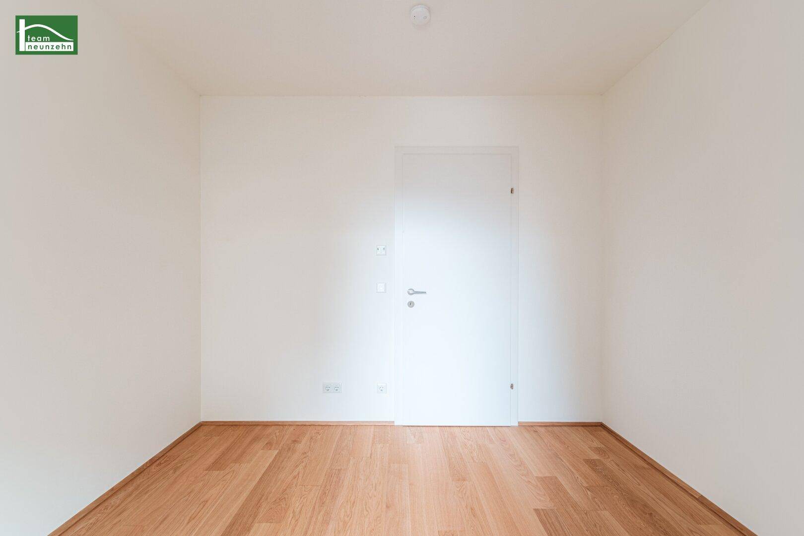 Unmöbliertes Zimmer mit hellem Holzboden und einer modernen weißen Tür.